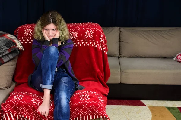 Horizontal concept photo of a young woman in a sweater and jeans sits on a chair in front of the camera in the home interior. Portrait of a girl with beautiful magnificent hair is missing on the sofa. — Stock Photo, Image