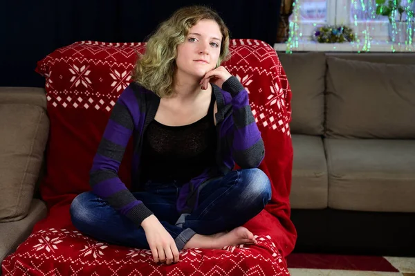 Πορτρέτο ενός κοριτσιού με όμορφα υπέροχα μαλλιά χαίρεται σε έναν καναπέ. Οριζόντια φωτογραφία έννοιας μιας νεαρής γυναίκας σε ένα πουλόβερ και τζιν κάθεται σε μια καρέκλα μπροστά από την κάμερα στο εσωτερικό του σπιτιού. — Φωτογραφία Αρχείου