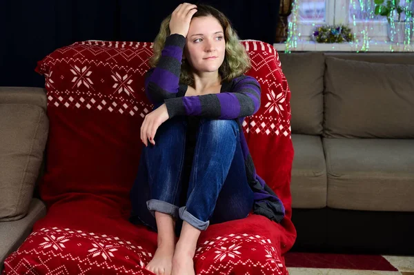 Porträtt av en flicka med vackert magnifikt hår glädjer sig på en soffa. Horisontellt konceptfoto av en ung kvinna i tröja och jeans sitter på en stol framför kameran i hemmets inredning. — Stockfoto