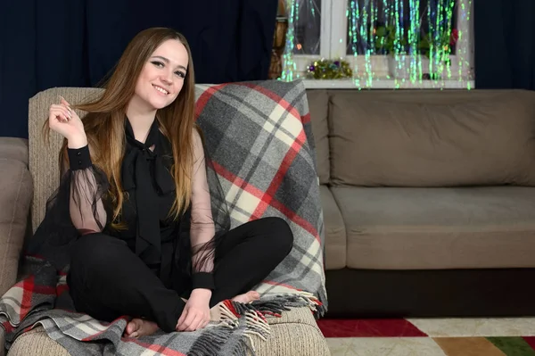 The concept of home comfort. Photo of pretty girl with excellent make-up in dark clothes brunette sitting on a sofa in the home interior, smiling.