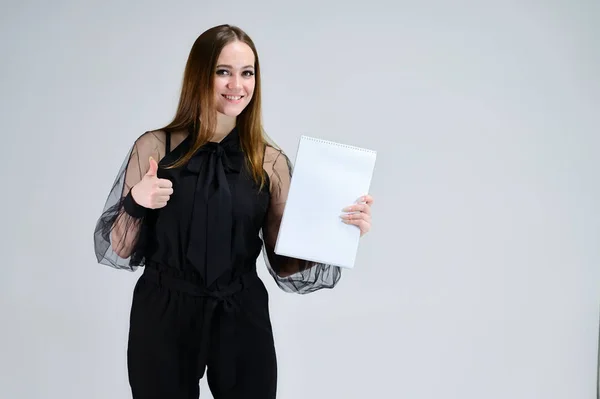 Retrato conceptual para una pancarta. Una foto de una chica morena bastante sonriente con una carpeta en sus manos con excelente maquillaje en ropa oscura está de pie sobre un fondo blanco . — Foto de Stock