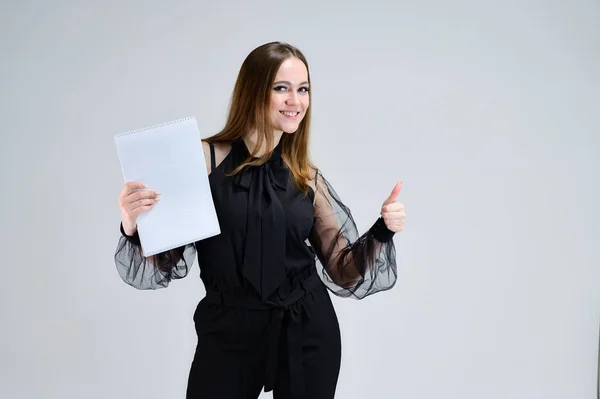 Retrato conceptual para una pancarta. Una foto de una chica morena bastante sonriente con una carpeta en sus manos con excelente maquillaje en ropa oscura está de pie sobre un fondo blanco . — Foto de Stock