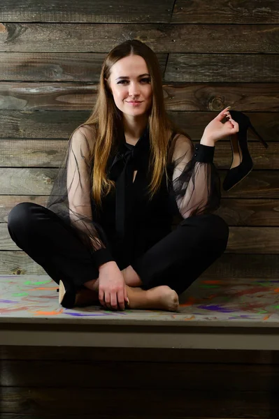 Het concept van een glamoureus portret. Een full-length foto van een schattige brunette met uitstekende make-up in donkere kleren zit op een stand op een houten achtergrond in het interieur. — Stockfoto