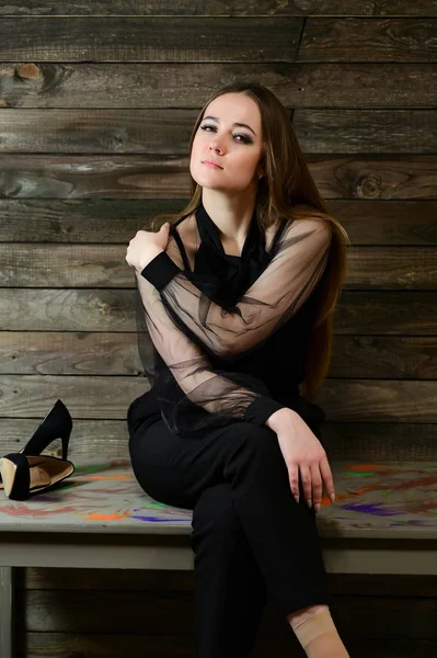 Het concept van een glamoureus portret. Een full-length foto van een schattige brunette met uitstekende make-up in donkere kleren zit op een stand op een houten achtergrond in het interieur. — Stockfoto