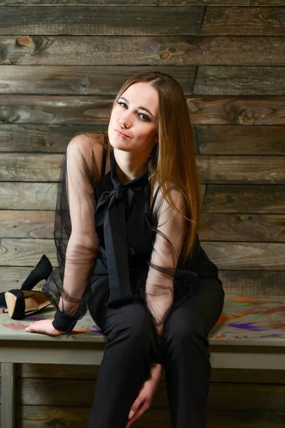 Η ιδέα ενός εντυπωσιακού πορτρέτου. Μια πλήρους μήκους φωτογραφία ενός χαριτωμένο μελαχρινή με εξαιρετικό μακιγιάζ σε σκούρα ρούχα κάθεται σε ένα περίπτερο σε ένα ξύλινο φόντο στο εσωτερικό του σπιτιού. — Φωτογραφία Αρχείου
