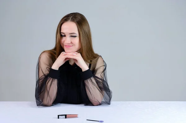 Kvinnan ler vackert mot kameran, visar känslor. Foto av en söt brunett med utmärkt makeup i mörka kläder sitter vid ett vitt bord på en vit bakgrund. — Stockfoto