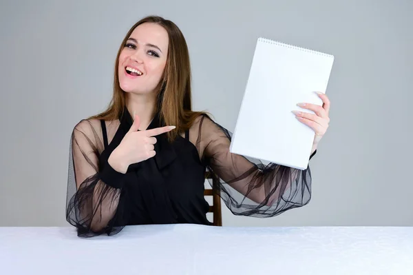 Una donna con una cartella in mano. Sorride meravigliosamente alla telecamera, si mostra con le mani. Foto di una bruna carina con grande trucco in abiti scuri seduta a un tavolo su uno sfondo bianco . — Foto Stock