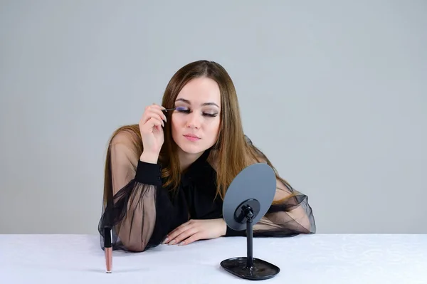 En kvinna sminkar sig framför en spegel. Foto av en söt brunett med stor makeup i mörka kläder sitter vid ett bord på en vit bakgrund. — Stockfoto