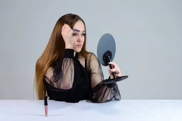En kvinna sminkar sig framför en spegel. Foto av en söt brunett med stor makeup i mörka kläder sitter vid ett bord på en vit bakgrund. — Stockfoto