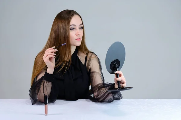En kvinna sminkar sig framför en spegel. Foto av en söt brunett med stor makeup i mörka kläder sitter vid ett bord på en vit bakgrund. — Stockfoto