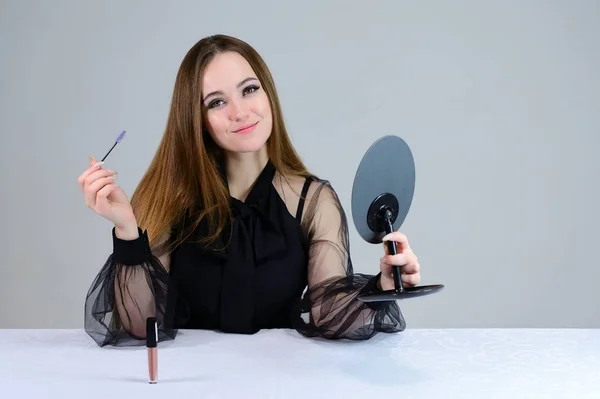 En kvinna sminkar sig framför en spegel. Foto av en söt brunett med stor makeup i mörka kläder sitter vid ett bord på en vit bakgrund. — Stockfoto