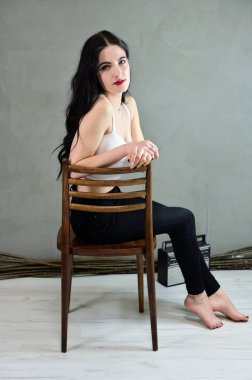 Göz alıcı bir kadın portresi konsepti. Uzun saçlı, güzel bir esmer kızın portresi. Kot pantolon ve gri arka planda bir tişört. Sandalyede oturuyor..