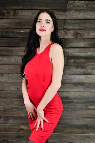 Retrato de una guapa morena con excelente maquillaje en un vestido rojo en un interior de madera se para frente a la cámara con una sonrisa. El concepto de un retrato femenino glamoroso . — Foto de Stock