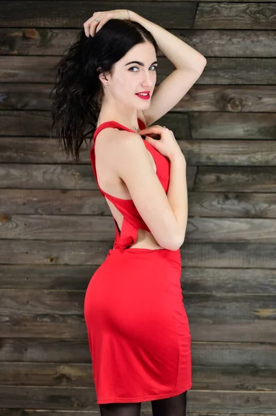 Portrait of a pretty brunette girl with excellent make-up in a red dress in a wooden interior stands in front of the camera with a smile. The concept of a glamorous female portrait. — Stock Photo, Image