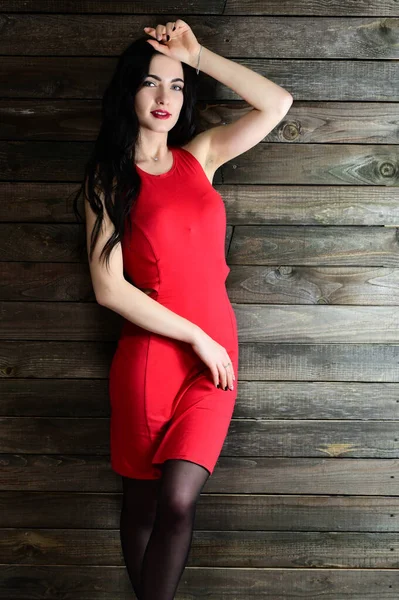 Koncepcja czarującego kobiecego portretu. Portret ładnej brunetki z doskonałym makijażem w czerwonej sukience w drewnianym wnętrzu stoi przed kamerą z uśmiechem. — Zdjęcie stockowe