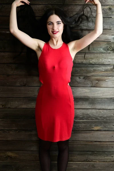 El concepto de un retrato femenino glamoroso. Retrato de una guapa morena de pelo largo con excelente maquillaje en un vestido rojo en un interior de madera está de pie delante de la cámara con una sonrisa . — Foto de Stock