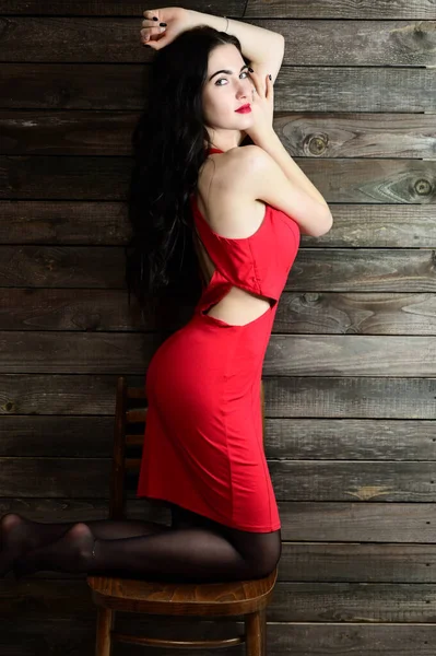O conceito de um retrato feminino glamouroso. Retrato de uma menina morena bonita com excelente maquiagem em um vestido vermelho em um interior de madeira fica na frente da câmera com um sorriso . — Fotografia de Stock