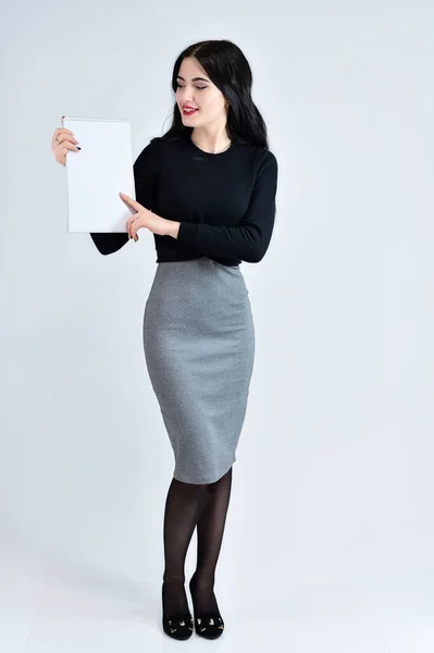 Una chica morena con una sonrisa de pelo largo con un maquillaje excelente en un vestido gris sobre un fondo blanco con una carpeta en las manos está de pie recta. Concepto de retrato de mujer de negocios . — Foto de Stock