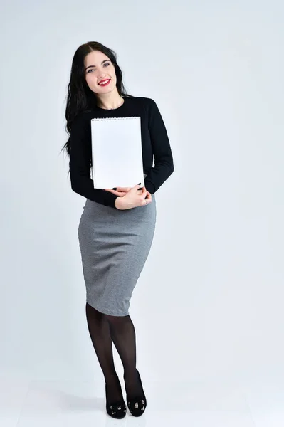 A brunette girl with a smile with long hair with excellent makeup in a gray dress on a white background with a folder in her hands is standing straight. Concept of business female photo portrait. — Stock Photo, Image