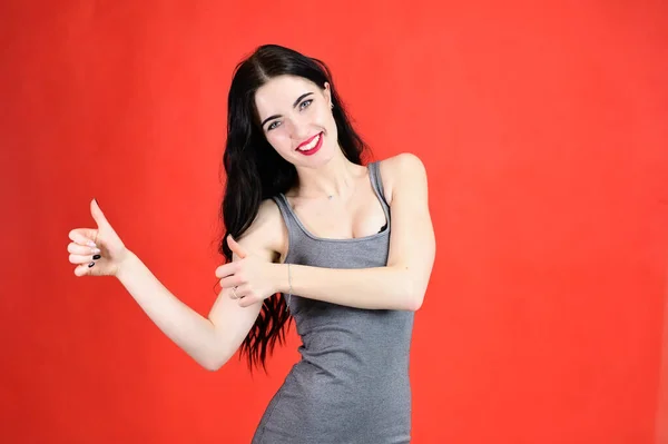 The concept of a beautiful female portrait with emotions. Portrait of a brunette girl with a smile with long hair with excellent makeup in a gray dress on a red background in different poses. — Stock Photo, Image