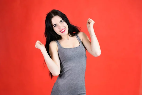 El concepto de un hermoso retrato femenino con emociones. Retrato de una chica morena con una sonrisa de pelo largo con excelente maquillaje en un vestido gris sobre un fondo rojo en diferentes poses . — Foto de Stock