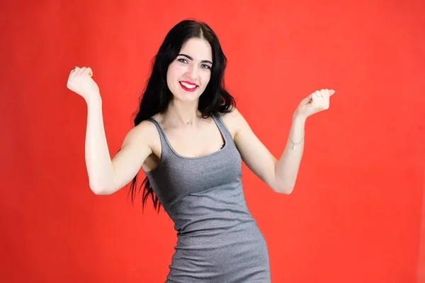 The concept of a beautiful female portrait with emotions. Portrait of a brunette girl with a smile with long hair with excellent makeup in a gray dress on a red background in different poses. — Stock Photo, Image