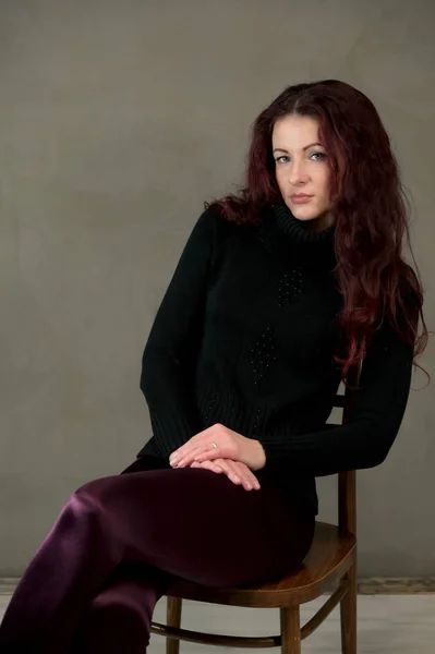 Ritratto di una bella donna bruna snella in un maglione scuro e con i capelli castani si siede su una sedia su uno sfondo grigio in studio. Il concetto di ritratto d'arte alla moda di un modello glamour — Foto Stock