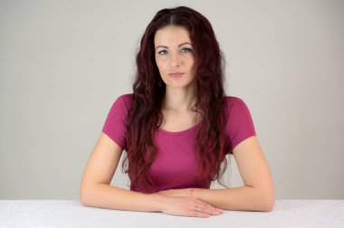 Model gülümsüyor ve poz vermekten ve konuşmaktan hoşlanıyor. Pembe elbiseli, kahverengi saçlı, ince, esmer bir kadının portresi stüdyoda beyaz bir arka planda bir masada oturuyor..