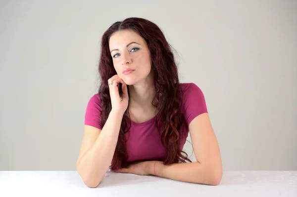 La modelo sonríe y disfruta posando y hablando. Retrato de una guapa morena delgada vestida de rosa y con el pelo castaño sentado en una mesa sobre un fondo blanco en el estudio . — Foto de Stock