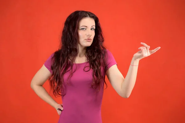 Il modello sta sorridendo e parlando carino alla fotocamera. Ritratto di una donna bruna piuttosto magra con emozioni in un vestito rosa e con i capelli castani su uno sfondo rosso in studio . — Foto Stock