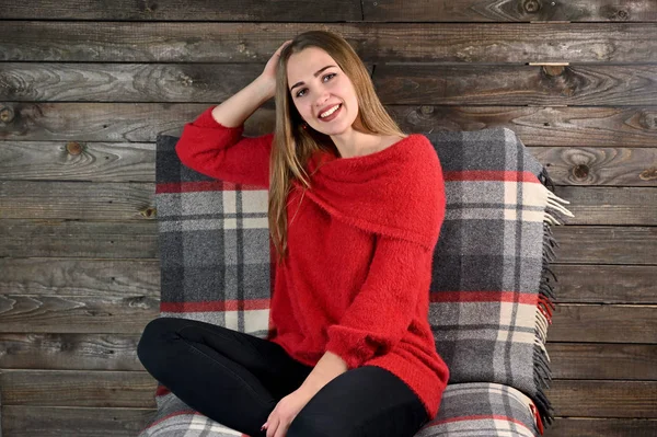 Koncepcja domowego komfortu. Zdjęcie ładnej dziewczyny z długimi włosami i doskonałym makijażem w czerwonym swetrze siedzącej na krześle na drewnianym tle w domowym wnętrzu. — Zdjęcie stockowe