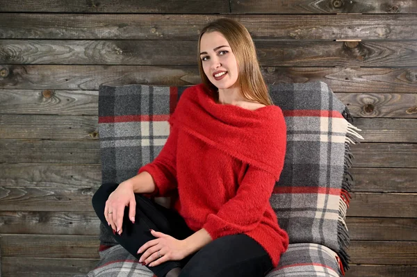 El concepto de confort en el hogar. Foto de una chica bonita con el pelo largo y excelente maquillaje en un suéter rojo sentado en una silla sobre un fondo de madera en el interior del hogar . — Foto de Stock