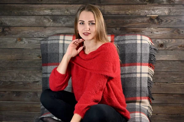 The concept of home comfort. Photo of a pretty girl with long hair and excellent make-up in a red sweater sitting in a chair on a wooden background in the home interior. — Stock Photo, Image