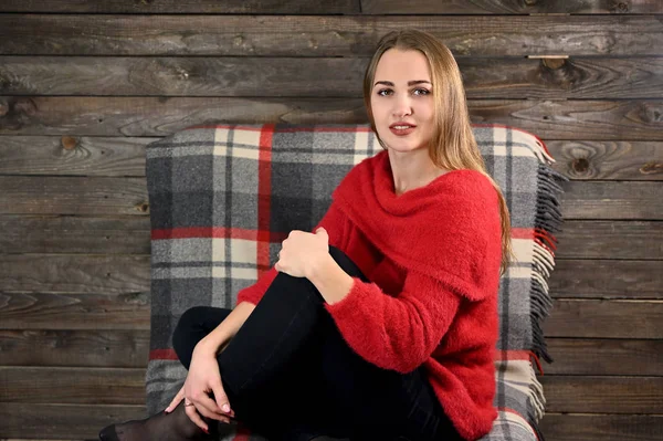 The concept of home comfort. Photo of a pretty girl with long hair and excellent make-up in a red sweater sitting in a chair on a wooden background in the home interior. — Stock Photo, Image