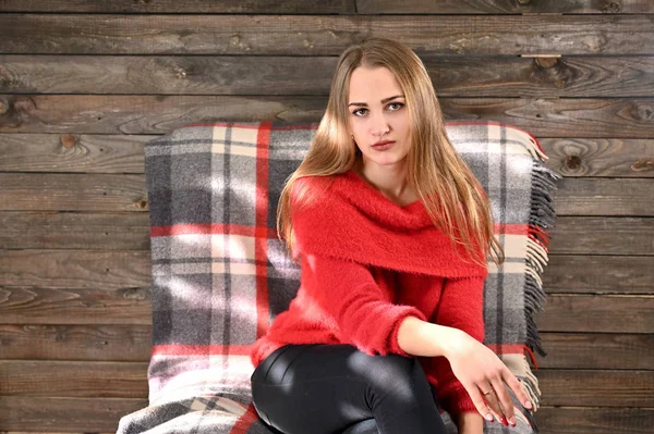 The concept of home comfort. Photo of a pretty girl with long hair and excellent make-up in a red sweater sitting bored in an armchair on a wooden background in the home interior in bright sunshine. — Stock Photo, Image