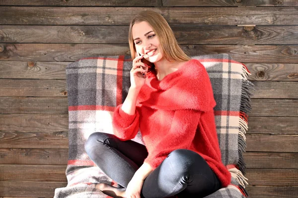 Photo of a pretty girl with long hair and excellent make-up in a red sweater sitting in a chair on a wooden background in the home interior uses a smartphone. The concept of home comfort. — Stock Photo, Image