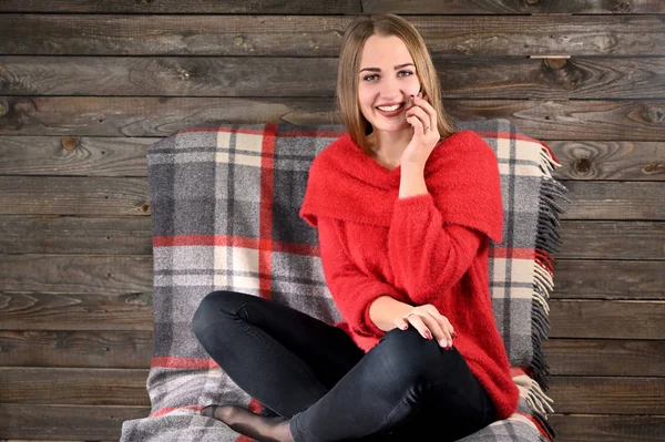 Zdjęcie ładnej dziewczyny z długimi włosami i doskonałym makijażem w czerwonym swetrze, siedzącej na krześle na drewnianym tle w domowym wnętrzu, korzysta ze smartfona. Koncepcja komfortu w domu. — Zdjęcie stockowe