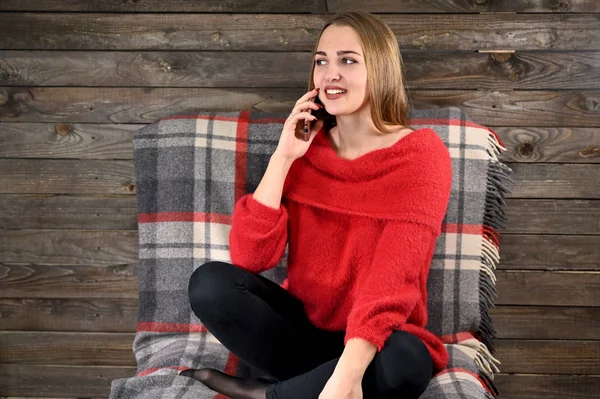 Photo of a pretty girl with long hair and excellent make-up in a red sweater sitting in a chair on a wooden background in the home interior uses a smartphone. The concept of home comfort. — Stock Photo, Image