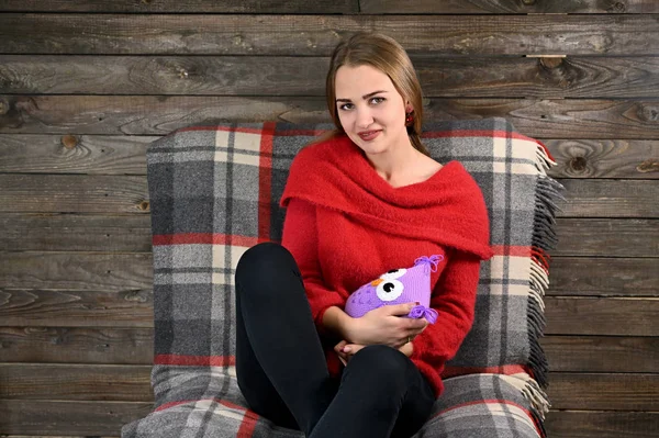 Foto de una chica bonita con el pelo largo y excelente maquillaje en un suéter rojo sentado en una silla sobre un fondo de madera en el interior del hogar con un juguete suave. El concepto de confort en el hogar . — Foto de Stock