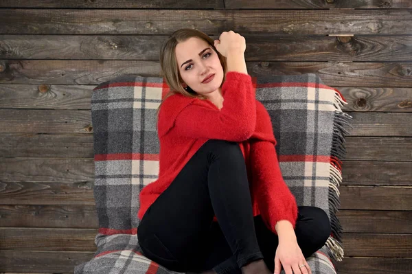 Zdjęcie ładnej dziewczyny z długimi włosami i doskonałym makijażem w czerwonym swetrze, siedzącej w fotelu na drewnianym tle w domowym wnętrzu. Koncepcja komfortu w domu. — Zdjęcie stockowe
