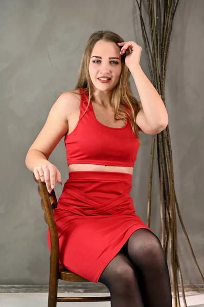 Foto vertical de uma menina muito sorridente com cabelos longos e excelente maquiagem em um vestido vermelho sentado em uma cadeira em várias poses. Retrato feminino conceito universal em um fundo cinza . — Fotografia de Stock