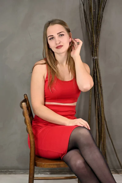 Вертикальне фото досить усміхненої дівчини з довгим волоссям і чудовим макіяжем у червоній сукні, що сидить на стільці в різних позах. Універсальний концепт жіночого портрета на сірому фоні . — стокове фото