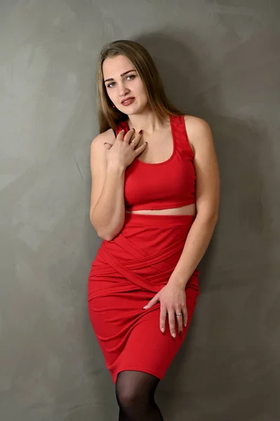 Das vertikale Foto eines hübschen lächelnden Mädchens mit langen Haaren und exzellentem Make-up in einem roten Kleid steht in verschiedenen Posen neben der Wand. Universelles Frauenporträt auf grauem Hintergrund. — Stockfoto