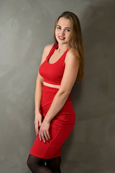 Vertical Photo of a pretty smiling girl with long hair and excellent make-up in a red dress stands next to the wall in various poses. Universal concept female portrait on a gray background. — 스톡 사진