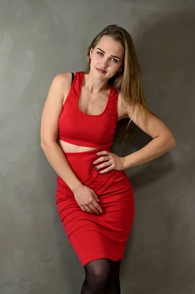 Verticaal Foto van een mooi lachend meisje met lang haar en uitstekende make-up in een rode jurk staat naast de muur in verschillende poses. Universeel concept vrouwelijk portret op een grijze achtergrond. — Stockfoto