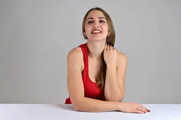 Universal koncept kvinnligt porträtt på en vit bakgrund. Horisontellt foto av en ganska leende flicka med långt hår och stor makeup sitter vid ett vitt bord i studion. — Stockfoto