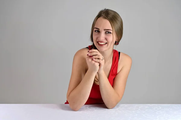 Horisontellt foto av en ganska leende flicka med långt hår och bra makeup sitter vid ett vitt bord i studion. Universal koncept kvinnligt porträtt på en vit bakgrund. — Stockfoto