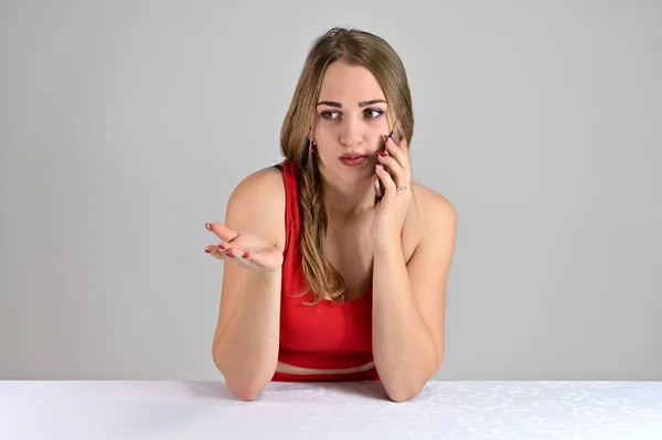 Horizontal photo of a pretty smiling girl with long hair and great makeup sitting at a white table in the studio using a telephone. Universal concept female portrait on a white background. — 스톡 사진