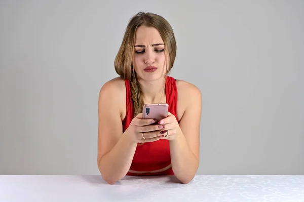 Horizontale foto van een mooi lachend meisje met lang haar en geweldige make-up zittend aan een witte tafel in de studio met behulp van een telefoon. Universeel concept vrouwelijk portret op een witte achtergrond. — Stockfoto