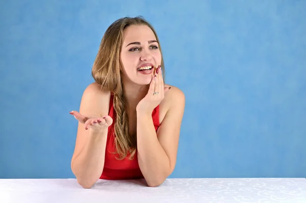 Retrato feminino conceito universal em um fundo azul. Foto horizontal de uma garota muito sorridente com cabelo comprido e ótima maquiagem sentada em uma mesa branca no estúdio . — Fotografia de Stock